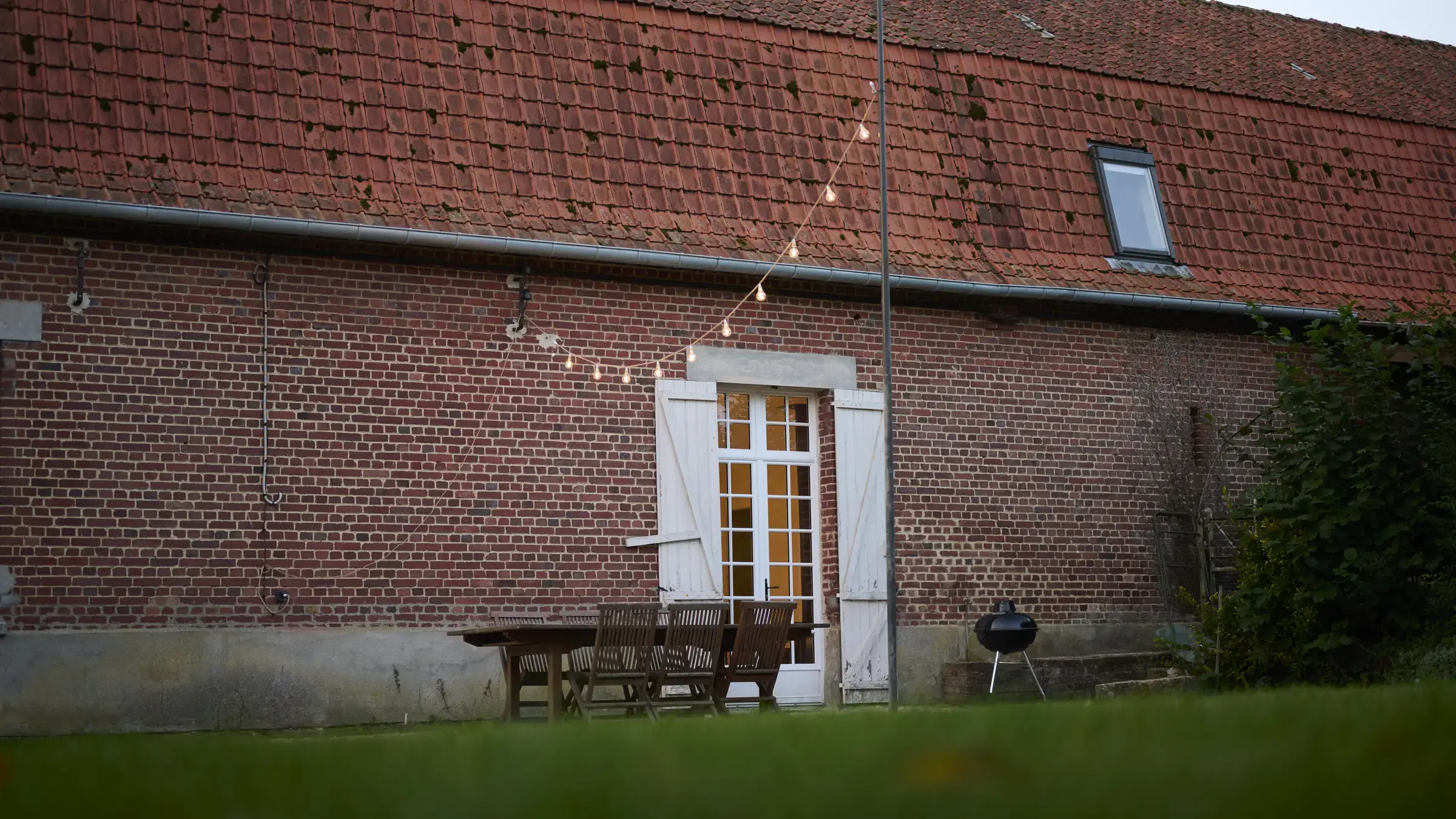 La façade en briques de la Maison de la Distillerie, sa porte vitrée, sa table de jardin et sa guirlande d’ampoules