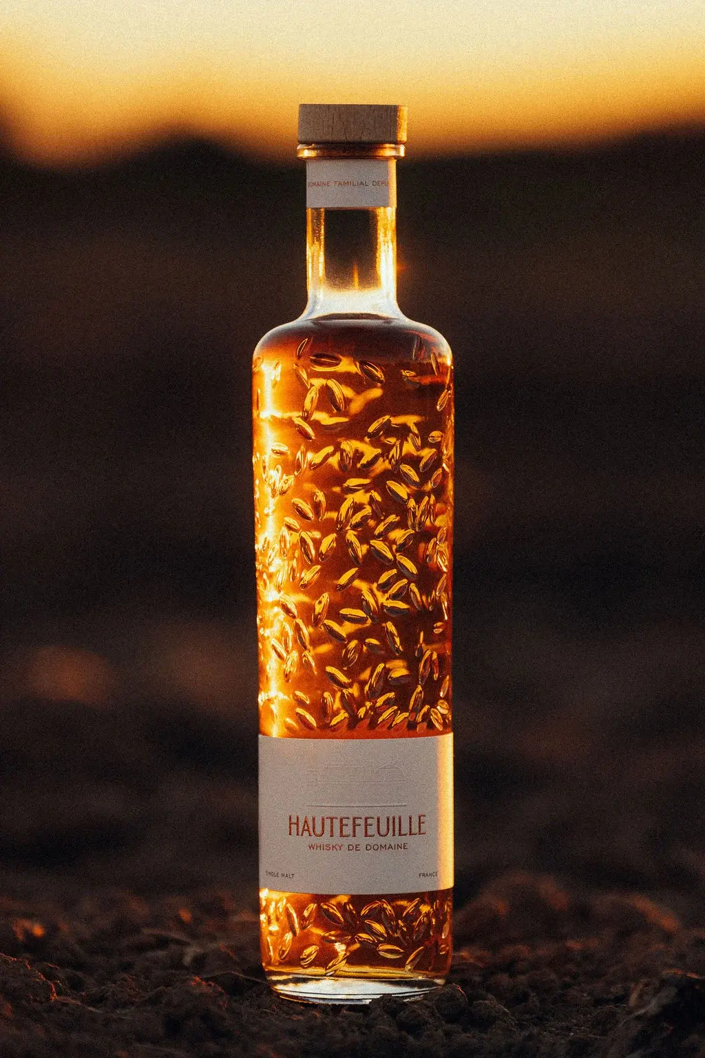Le flacon du Whisky de Domaine posé sur la terre d’un champ au soleil couchant : bouchon de bois, verre gaufré de grains d’orge, étiquette crème